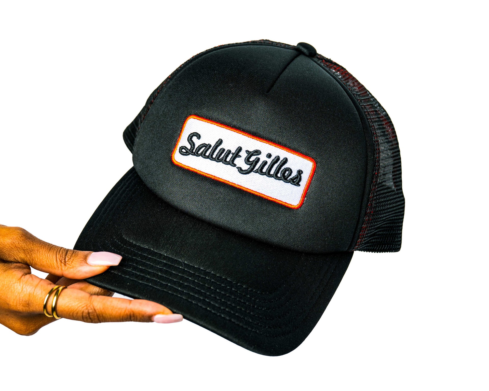 Main tenant une casquette trucker noire avec patch brodé ‘Salut Gilles’, vue de côté.